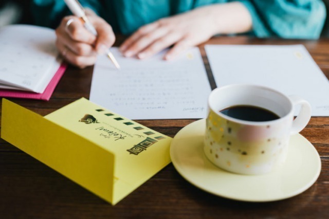 テーブルで手紙を書いている女性。コヒーカップと黄色い封筒。